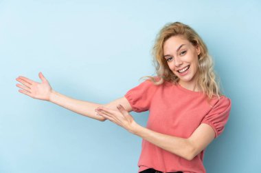 Mavi arka planda izole edilmiş genç sarışın kadın davet etmek için ellerini yana uzatıyor.