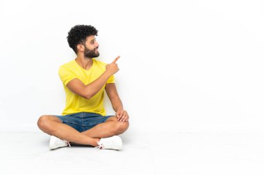 Genç Faslı yakışıklı adam izole arka planda yerde oturuyor ve işaret parmağıyla işaret ediyor.