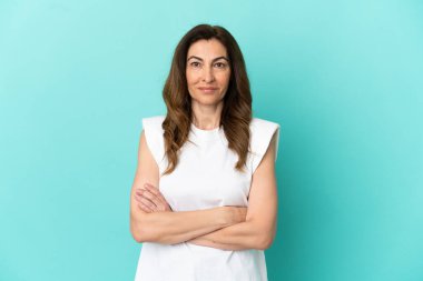 Orta yaşlı, beyaz bir kadın, mavi arka planda izole edilmiş, kollarını ön cephede kavuşturuyor.