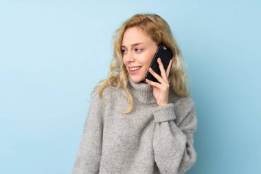 Mavi arka planda izole edilmiş süveter giyen genç sarışın bir kadın cep telefonuyla sohbet ediyor.