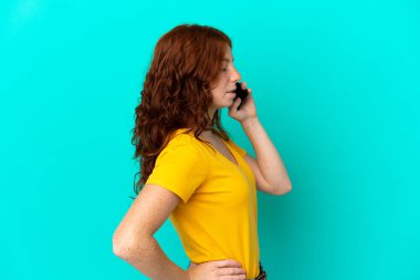 Cep telefonu kullanan genç kızılımsı bir kadın, mavi arka planda izole edilmiş, sırt ağrısı çekiyor, gayret gösterdiği için.