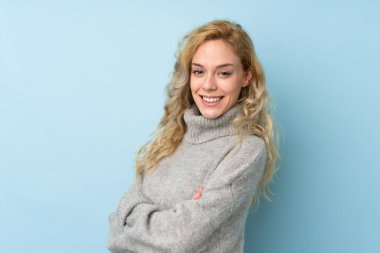 Mavi arka planda izole edilmiş bir süveter giyen genç sarışın kadın kollarını kavuşturup ileriye bakıyor.
