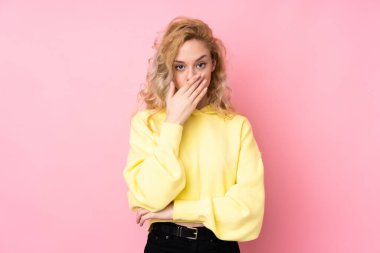 Pembe arka planda izole edilmiş eşofman üstü giyen genç sarışın kadın elleriyle ağzını kapatıyor.