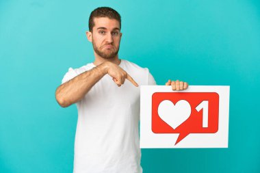 Yakışıklı sarışın, izole edilmiş mavi arkaplan üzerinde ikon gibi bir pankart tutan ve onu işaret eden.