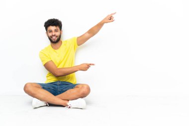 Genç Faslı yakışıklı adam, izole edilmiş arka planda yerde oturuyor. Parmaklarını kenarı gösteriyor ve bir ürün sunuyor.