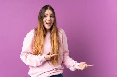 Mor arka planda izole edilmiş genç beyaz kadın şaşırmış ve işaret ediyor.