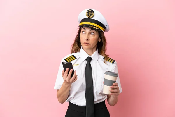 Airplane middle aged pilot woman isolated on pink background holding coffee to take away and a mobile while thinking something