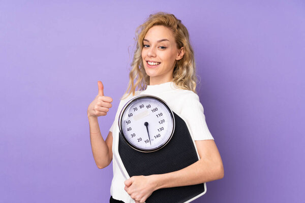 Young blonde woman isolated on purple background holding weighing machine with thumb up