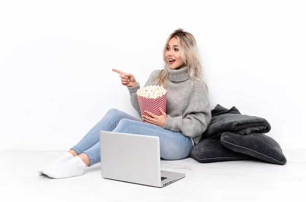 年轻的金发女孩一边吃爆米花 一边在笔记本电脑上看电影 指指点点旁边 展示自己的产品 — 图库照片