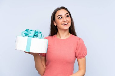 Pasta şefi mavi arka planda izole edilmiş büyük bir pasta tutuyor gülümserken yukarı bakıyor.