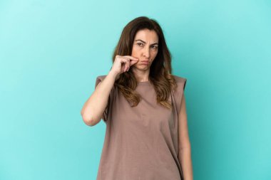 Mavi arka planda izole edilmiş orta yaşlı beyaz kadın sessizlik işareti gösteriyor.