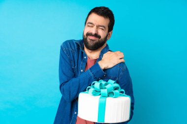 İzole edilmiş mavi arka planda büyük bir pastası olan genç yakışıklı adam çabaladığı için omzunda acıdan muzdarip.