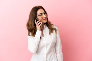 Pembe arka planda izole edilmiş orta yaşlı beyaz kadın cep telefonuyla sohbet ediyor.