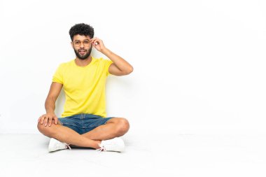 Genç Faslı yakışıklı, gözlüklü ve şaşırmış bir şekilde izole bir zeminde oturuyor.