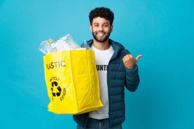 Elinde plastik şişelerle dolu bir çanta tutan genç Faslı bir adam izole edilmiş arka planı geri dönüştürmek için bir ürün sunmak için kenarı işaret ediyor.
