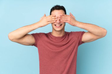 Beyaz tenli yakışıklı adam mavi arka planda izole edilmiş gözleri elleriyle kapatıyor.