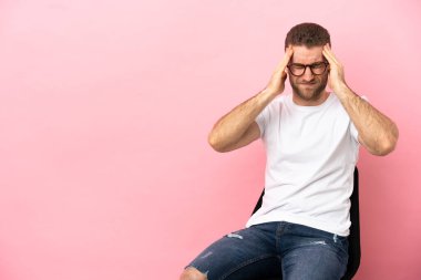 Genç adam, izole edilmiş pembe arka planda, baş ağrısı olan bir sandalyede oturuyor.