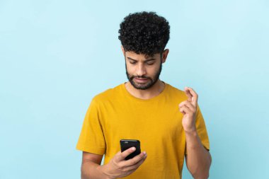 Faslı genç adam mavi arka planda parmakları çapraz cep telefonu kullanarak izole edildi.