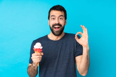 Kornet dondurması olan genç bir adam izole edilmiş mavi arka planda şaşırmış ve iyi bir işaret gösteriyor.