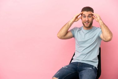 İzole edilmiş pembe arka planda bir sandalyede oturan genç adam sürpriz bir ifadeyle