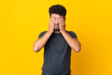 Faslı genç bir adam sarı arka planda izole edilmiş. Gözlerini elleriyle kapatıyor.