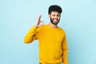 Faslı genç adam mavi arka planda izole edilmiş. Parmaklarıyla işaret veriyor.