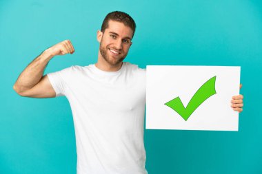 Yakışıklı sarışın adam izole edilmiş mavi arka planda elinde yeşil işaret simgesi olan bir pankart tutuyor ve güçlü bir hareket yapıyor.