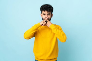 Mavi arka planda izole edilmiş genç Faslı bir adam sessizlik işareti gösteriyor.
