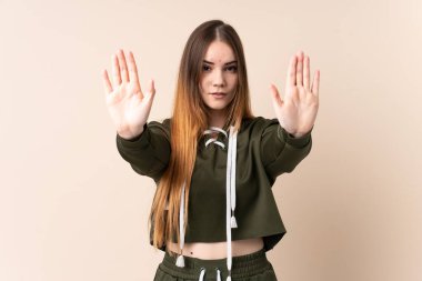 Beyaz tenli genç bir kadın bej arka planda izole edilmiş. Durma hareketi yapıyor ve hayal kırıklığına uğruyor.