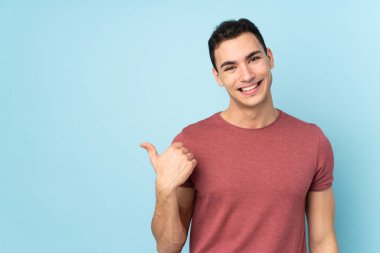 Mavi arka planda izole edilmiş beyaz, yakışıklı genç bir adam bir ürün sunmak için kenarı işaret ediyor.