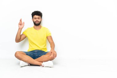 Genç Faslı yakışıklı adam izole bir zeminde oturuyor. Parmakları çapraz ve en iyisini diliyor.