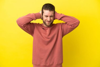 Sarışın yakışıklı adam izole edilmiş sarı arka planda hayal kırıklığına uğramış ve kulaklarını kapatmış.