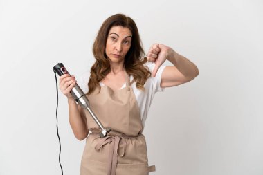 Beyaz arka planda izole edilmiş el blenderi kullanan orta yaşlı beyaz kadın, başparmağın olumsuz ifadesini gösteriyor.