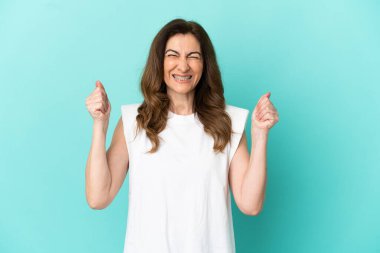 Mavi arka planda izole edilmiş orta yaşlı beyaz kadın galibiyeti kutluyor.