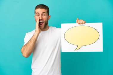 Yakışıklı sarışın, izole edilmiş mavi arkaplan üzerinde konuşma balonu ikonu olan bir levha tutuyor ve bağırıyor.