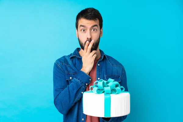 Young handsome man with a big cake over isolated blue background surprised and shocked while looking right