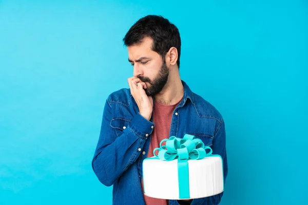 Young handsome man with a big cake over isolated blue background having doubts