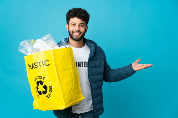 Young Moroccan man holding a bag full of plastic bottles to recycle over isolated background extending hands to the side for inviting to come