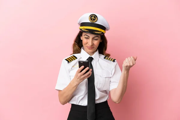 Airplane middle aged pilot woman isolated on pink background with phone in victory position
