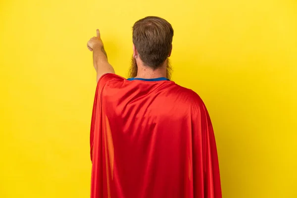 レッドヘッドスーパーヒーロー男は インデックス指で後ろを指して黄色の背景に隔離 — ストック写真