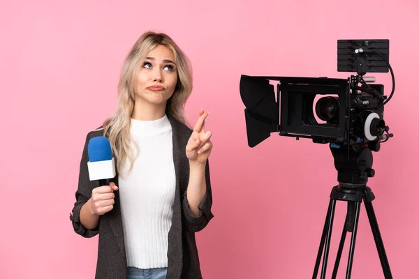 Reporter woman holding a microphone and reporting news over isolated pink background with fingers crossing and wishing the best