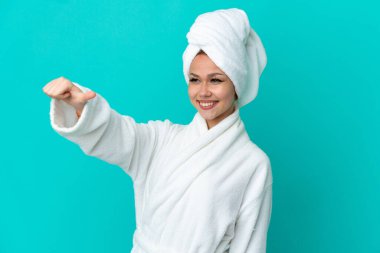 İzole edilmiş mavi arka planda bornozlu genç sarışın kız baş parmak işareti yapıyor.