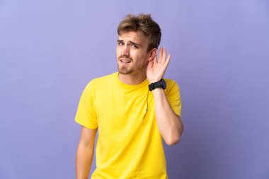 Mor arka planda izole edilmiş genç, yakışıklı sarışın bir adam. Kulağına elini sürerek bir şeyler dinliyor.
