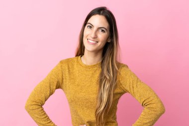 Pembe arka planda izole edilmiş genç beyaz kadın kolları kalçasında ve gülümsüyor.