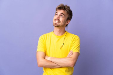 Genç yakışıklı sarışın adam mor arka planda izole edilmiş gülümserken yukarı bakıyor.