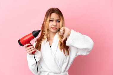 Genç sarışın kız elinde saç kurutma makinesiyle izole edilmiş pembe arka planda negatif bir ifadeyle baş parmağını gösteriyor.