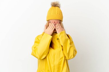 Yağmura dayanıklı bir ceket giyen genç sarışın kız izole edilmiş beyaz arka plan, gözleri elleriyle kaplıyor.