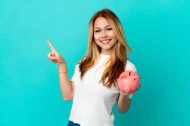 İzole edilmiş mavi arka planda kumbarayı elinde tutan genç sarışın kız parmağını yana uzattı ve bir ürün sundu.