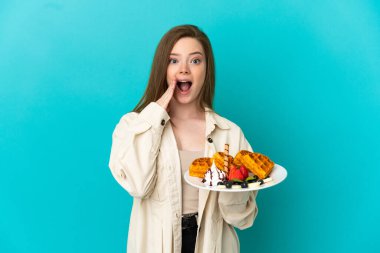 İzole edilmiş mavi arka planda waffle tutan genç kız şaşırmış ve şok olmuş yüz ifadesi ile