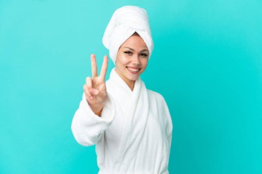 Mavi arka plan üzerinde bornozlu genç sarışın kız gülümsüyor ve zafer işareti gösteriyor.